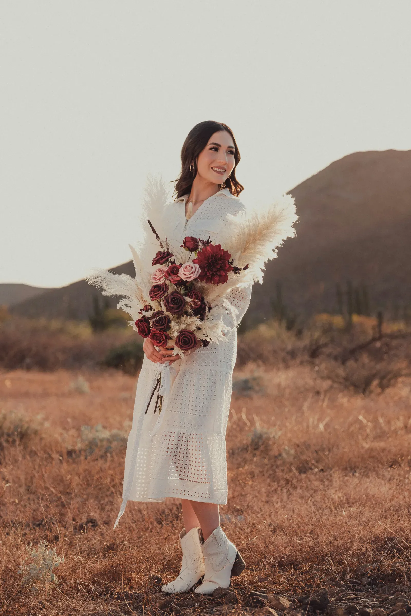The bride stands in her white dress with a pampas-and-rose bouquet, golden hills rolling away behind her.