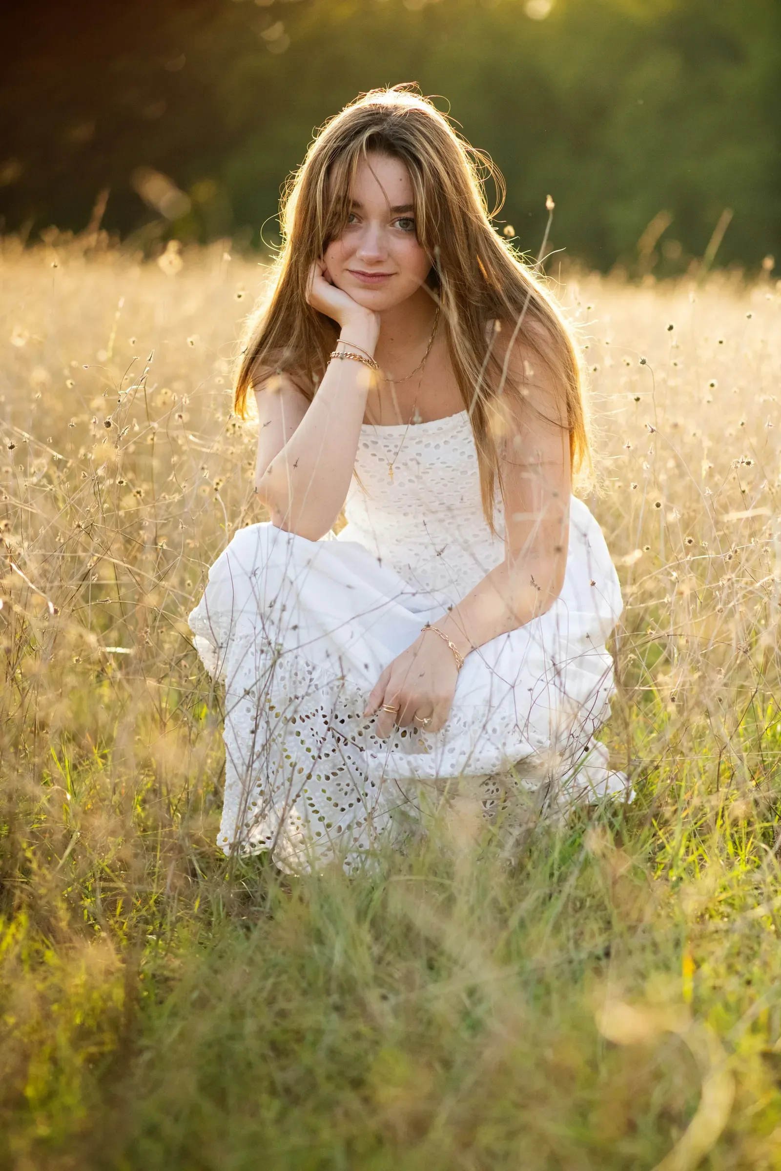 Knee-deep in backlit September grass in a white dress, hand at her chin.