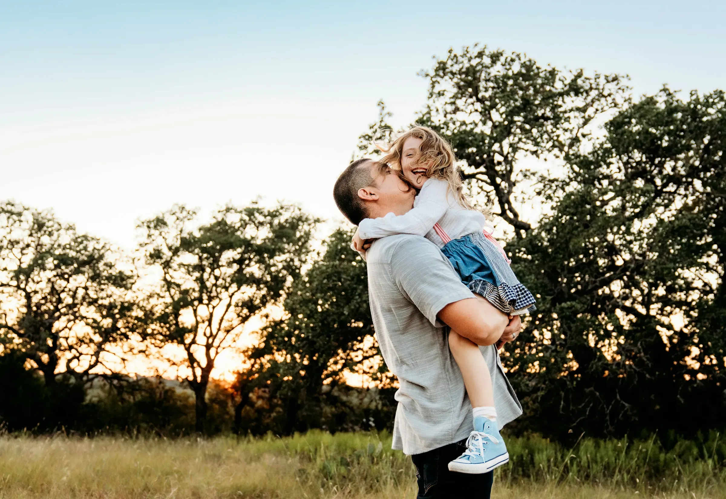 Dad scoops his daughter up for a kiss, backlit by the low sun.