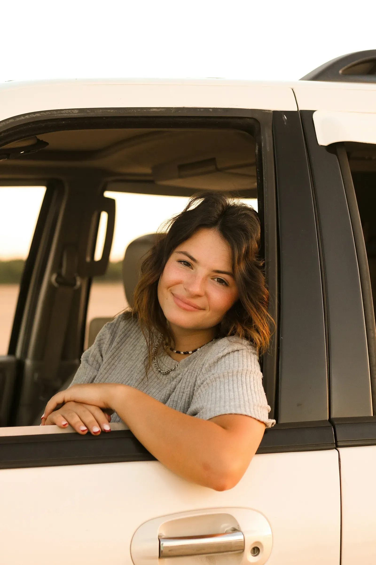 Leaning out the driver's window with her arms folded, easy smile, sunset on the water.