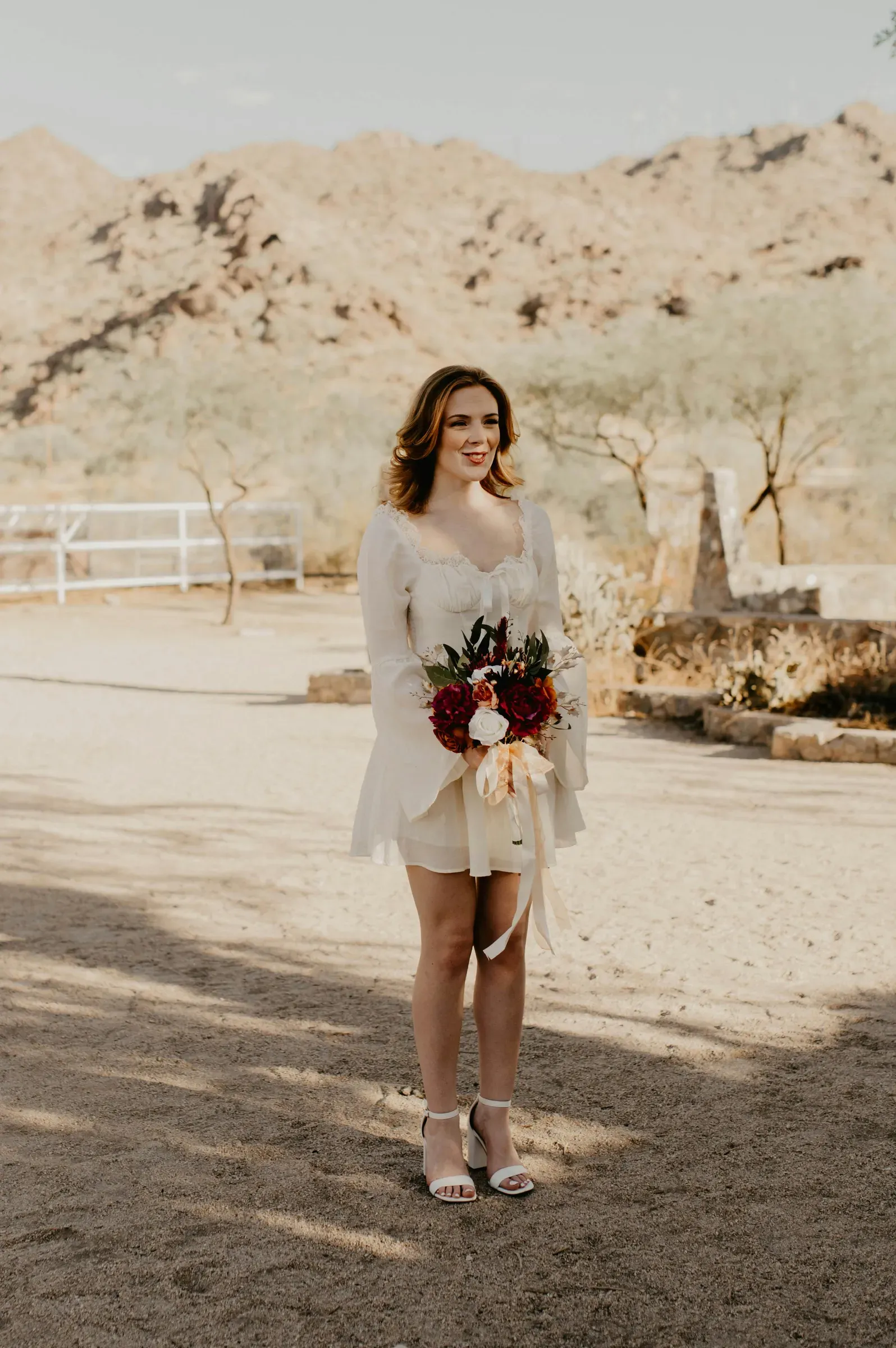The bride in her short white dress holds her rust-and-cream bouquet by the corral fence, desert hillside behind.