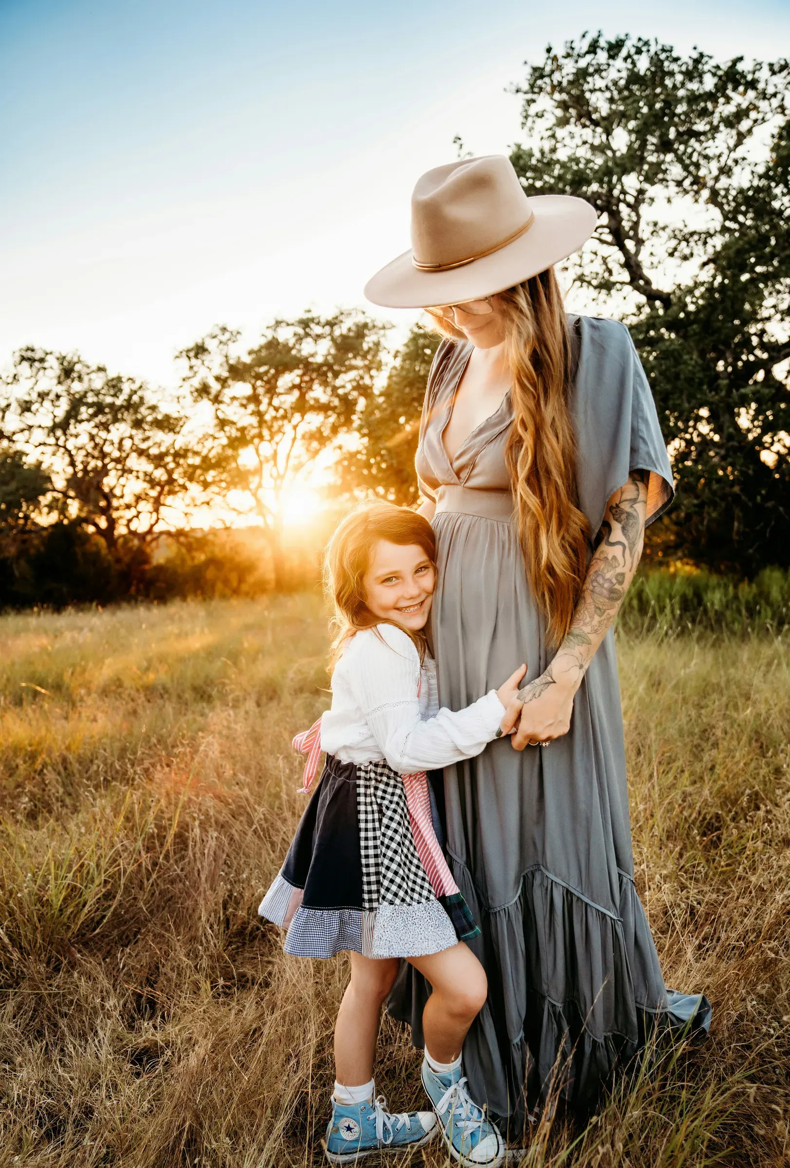 The girl leans into her mom's long dress while the sun drops behind the trees.