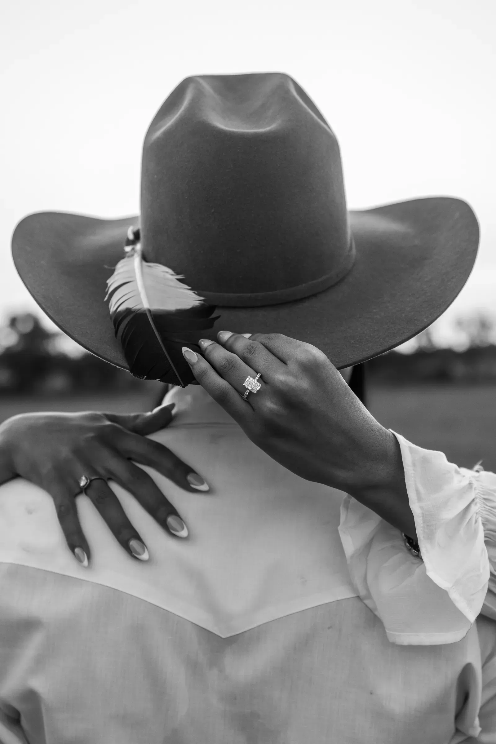 A black-and-white detail over his shoulder — her ring hand, a feather tucked in the hat band.