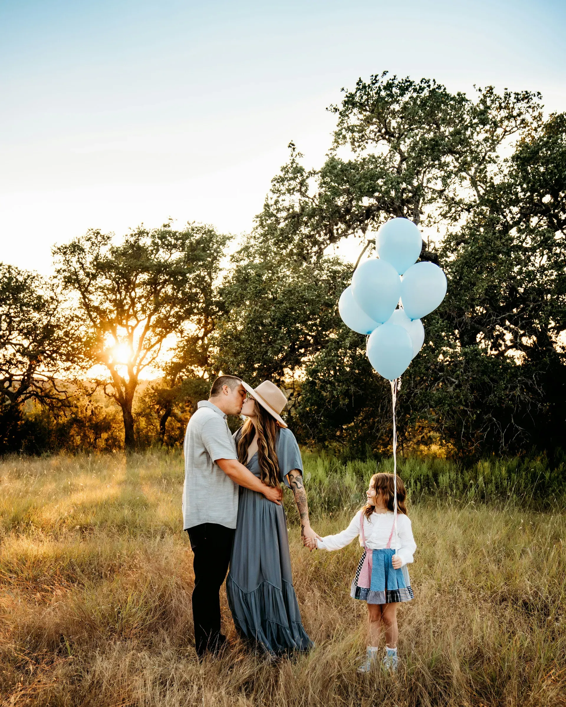 Her parents kiss behind her while she holds the balloons at golden hour.