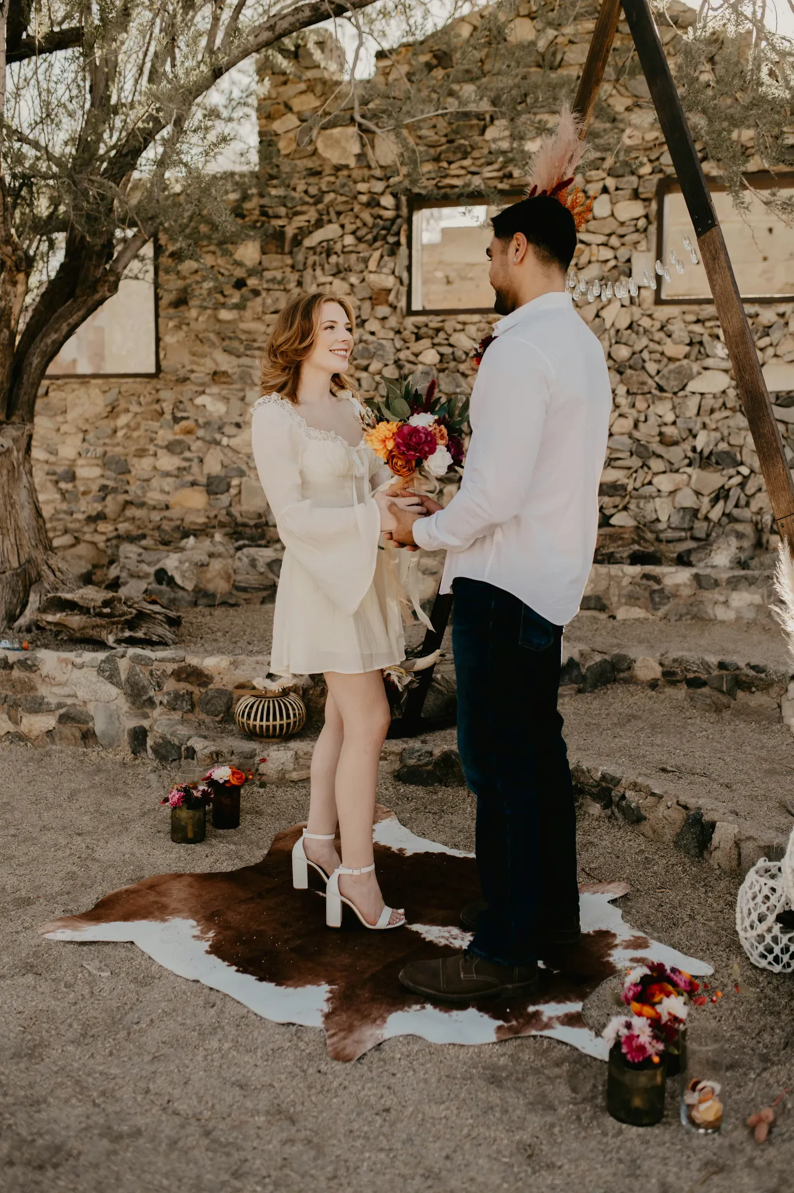 The couple pauses together under a bare desert tree, the bouquet still in her hand.